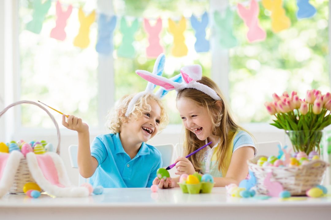 Kinder mit Hasenohren bemalen Ostereier gemeinsam und lachen an einem festlich dekorierten Tisch