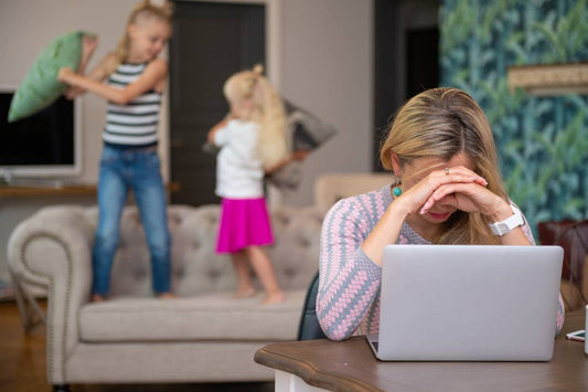 Müde Mama total erschöpft vor dem Laptop. Im Hintergrund stehen zwei Mädchen auf der Couch und machen eine Kissenschlacht