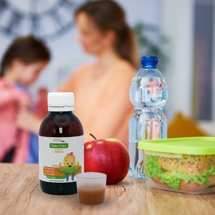Hokus Fokus Sirup mit Dosierbecher steht neben einem Apfel, einer Wasserflasche und einer Brotdose, im Hintergrund hilft eine Mutter ihrer Tochter beim Jacke anziehen